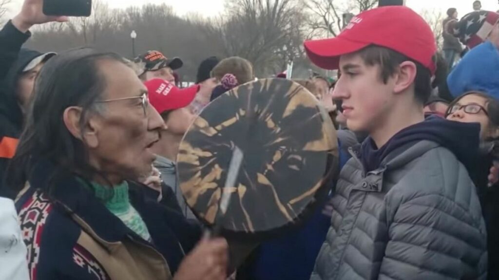Nick Sandmann wears a black coat and a red face cap while talking to a man on glasses