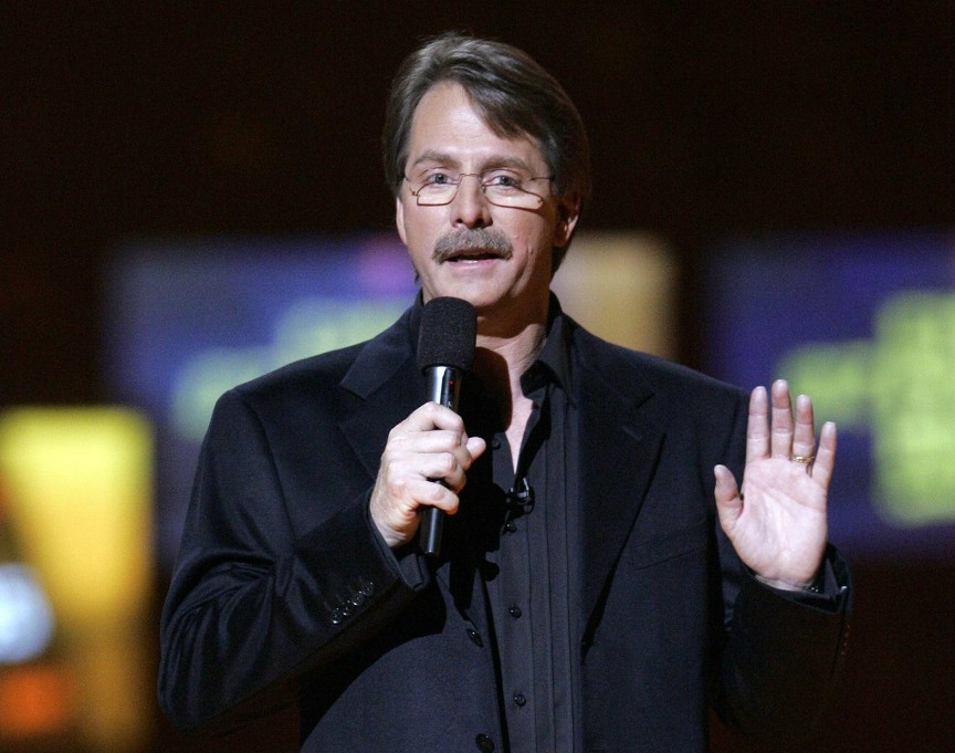 Jeff Foxworthy wearing a black suit on stage with glasses on his face